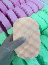 Load image into Gallery viewer, A gloved hand holding a colourful handmade exfoliating soap sponge with fresh batches of sponges lined up in rows in the background.
